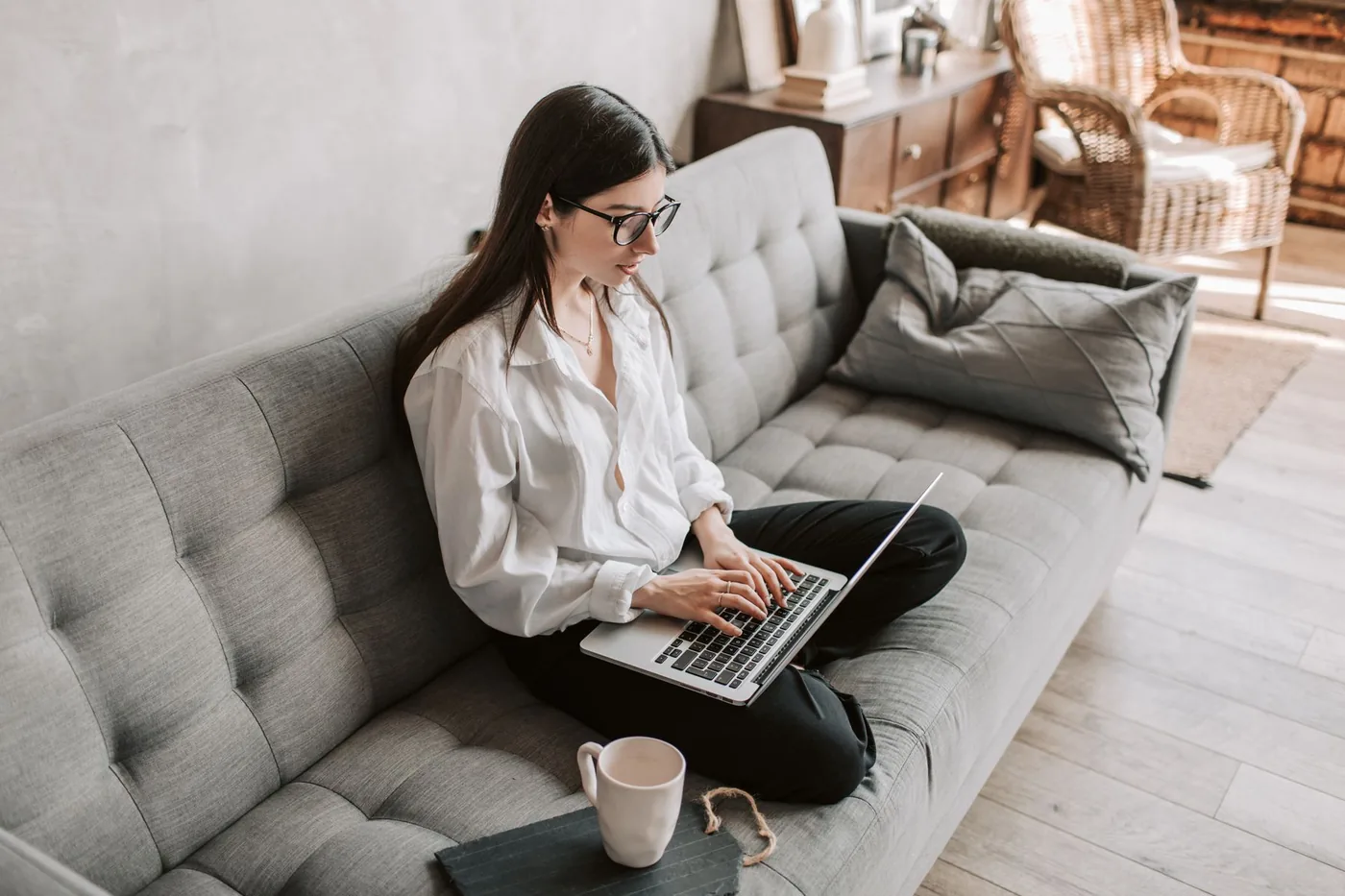 An adult using a laptop on a grey sofa beside an open notebook.