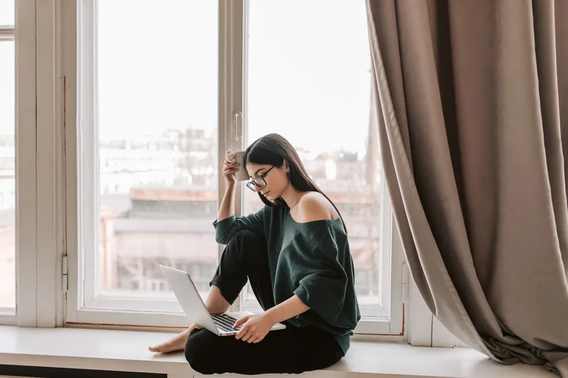 An adult writing in a notebook beside a window with a laptop nearby.