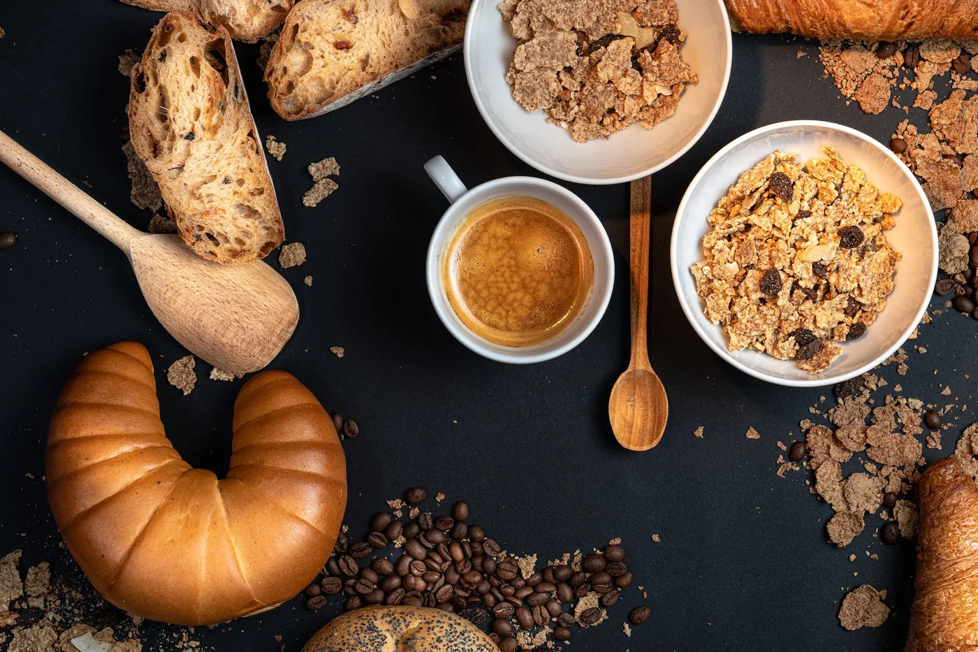 Bread, croissants, cereal bowls and a cup of coffee arranged on a dark table.