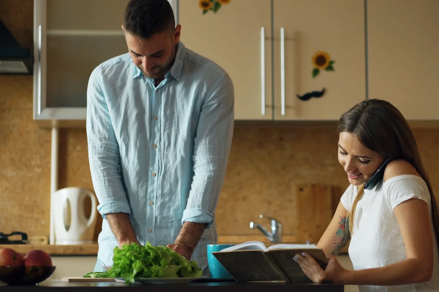 Two adults preparing food together beside an open recipe book in a kitchen.