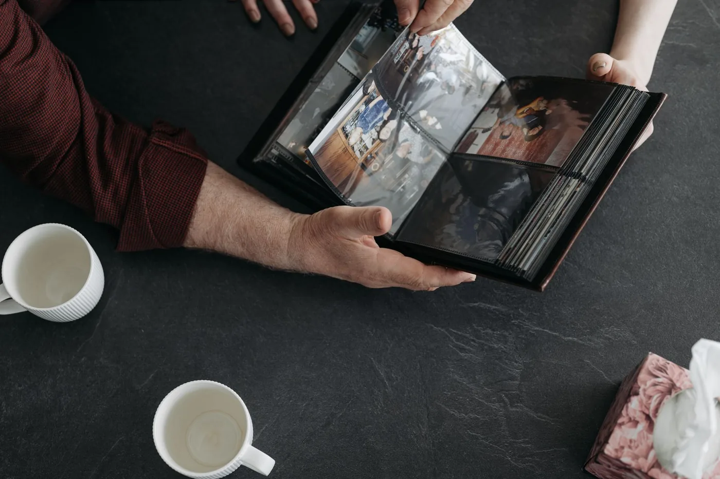 Hands holding an open photo album beside cups of coffee on a table.